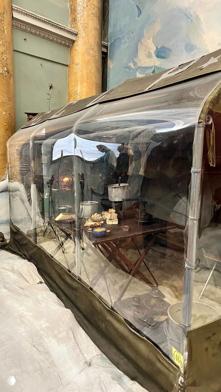 Крупный план прозрачной части палатки-витрины: внутри котелки, посуда и складная мебель экспедиционного быта, отражения посетителей.