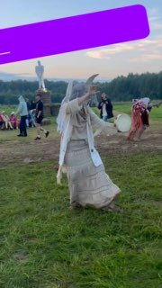 Фигура в длинном белом костюме с барабаном на открытой поляне Архстояния, люди и художественные объекты на заднем плане в дневном свете.