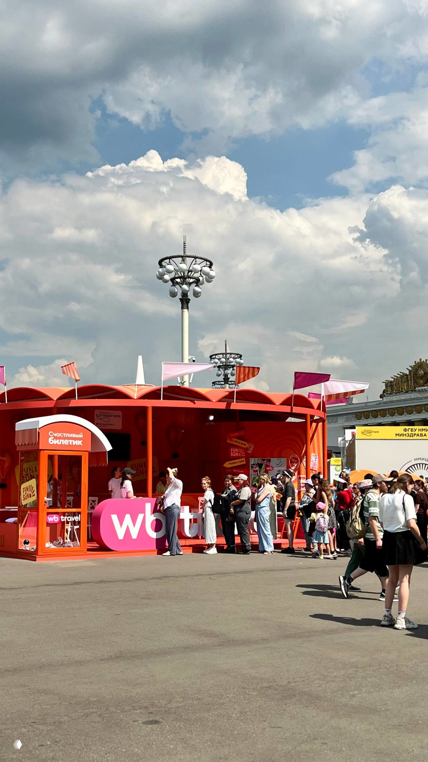Оранжевый стенд WB Travel и очередь посетителей у промо-точки на яркой летней площадке ВДНХ, люди в ожидании информации и сувениров.