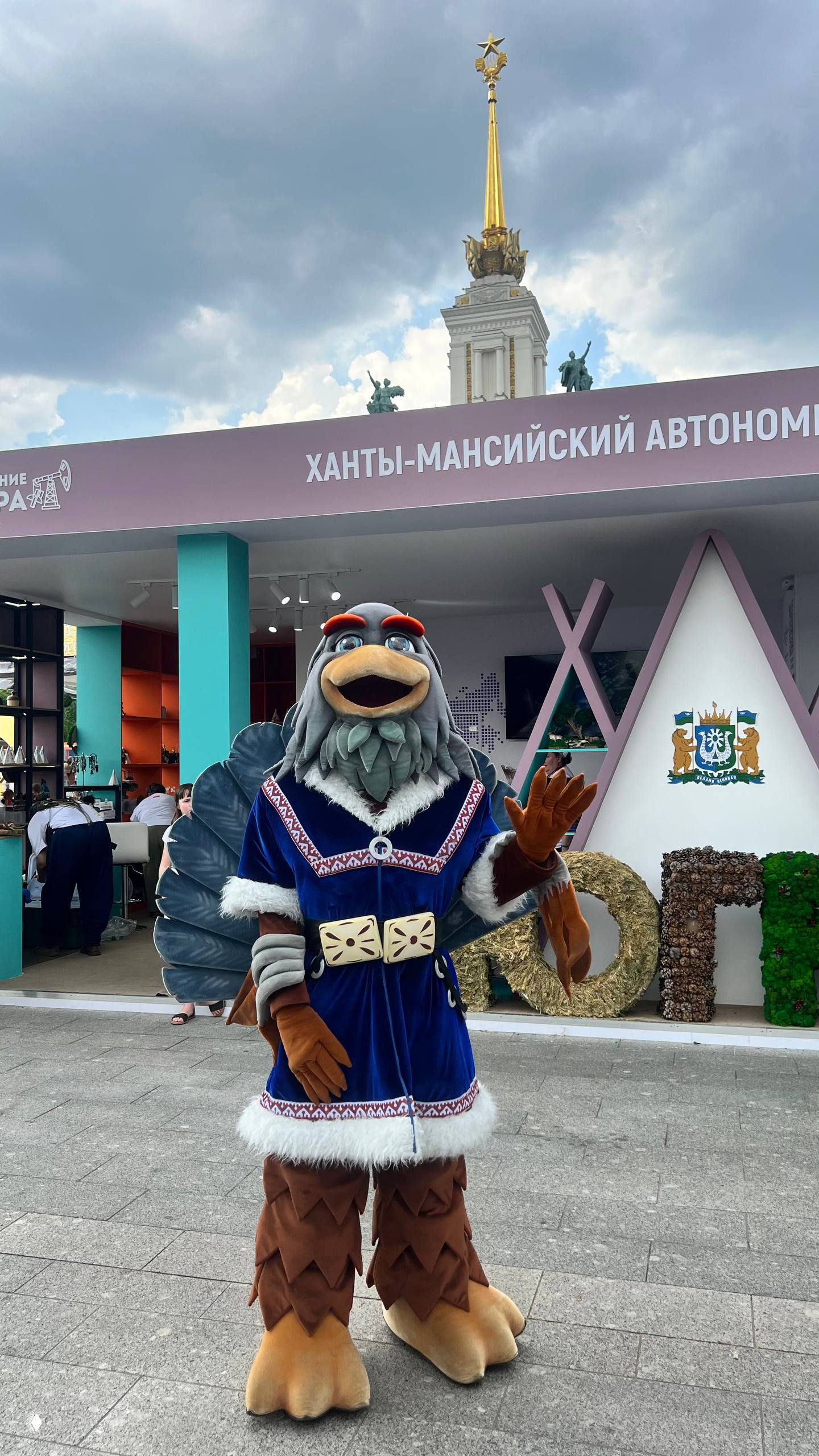 Маскот в костюме птицы у стенда Ханты‑Мансийского автономного округа: ростовая кукла, элементы народного костюма и фон экспозиции.