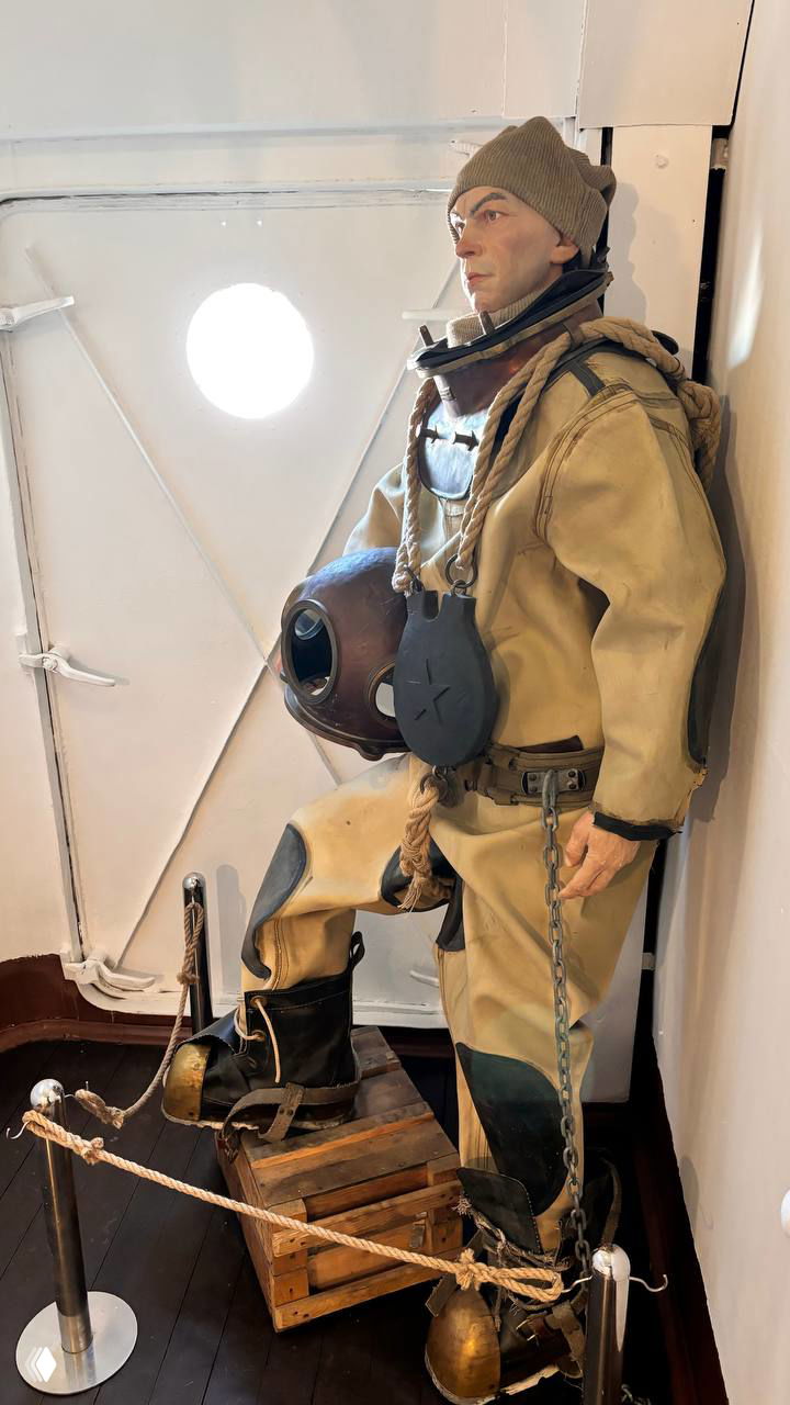 Экспозиция исторического водолаза в костюме: манекен в тяжёлом снаряжении, с шлемом и грузами, выставка по истории спасательных работ на корабле.