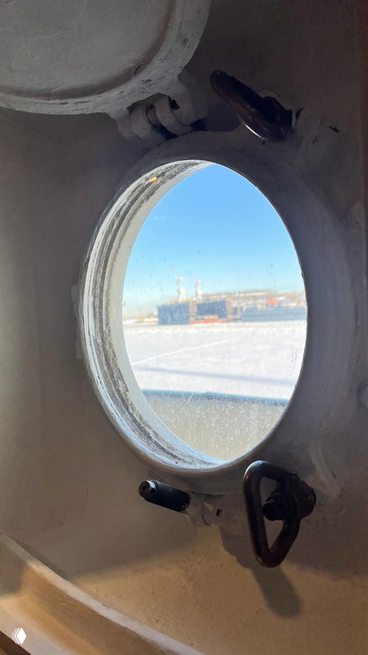 Вид через иллюминатор на ледовую поверхность и другой пришвартованный корабль: рамка окна, потёртая краска и арктический пейзаж за стеклом.