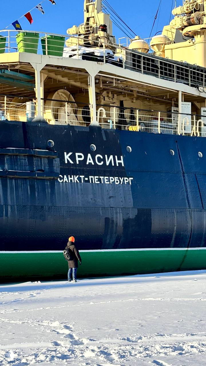 Набережная, на которой стоит ледокол «Красин»: крупный план борта с надписью КРАСИН САНКТ-ПЕТЕРБУРГ, человек в оранжевой шапке для масштаба.