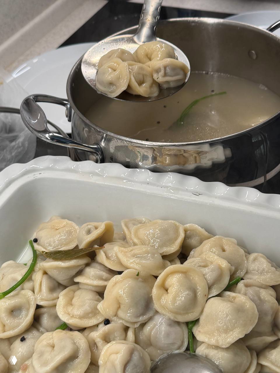 Отваренные пельмени в белой сервировочной посуде и шумовка над кастрюлей с бульоном, пельмени уже подаются к столу со сметаной и зеленью.