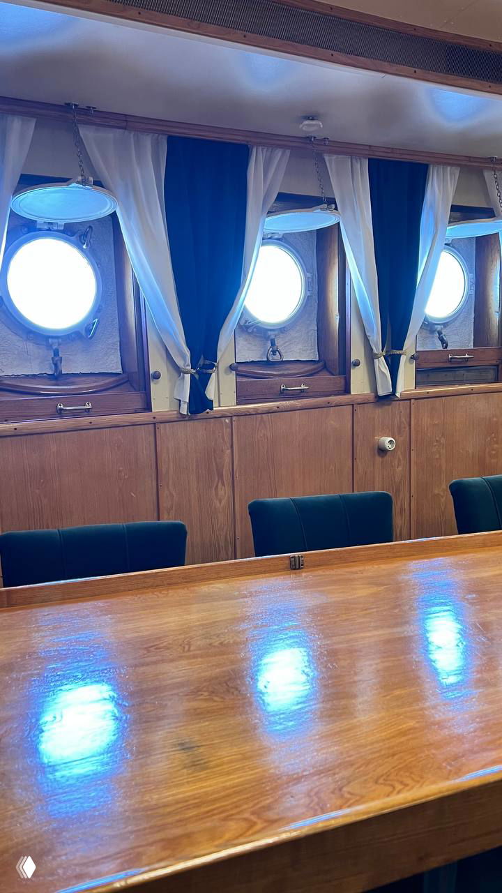 Каюта с круглым иллюминатором и столовой зоной: деревянная отделка, занавески и зеленые стулья, интерьер офицерской комнаты на «Красине».