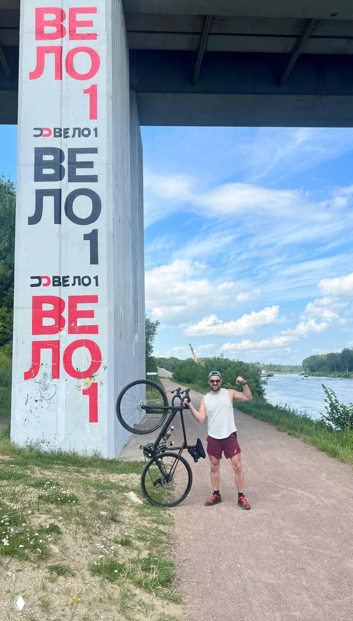Райдер держит велосипед у бетонной опоры с надписью «ВЕЛО1» на фоне реки и открытого неба — одиночная спортивная сцена на набережной.