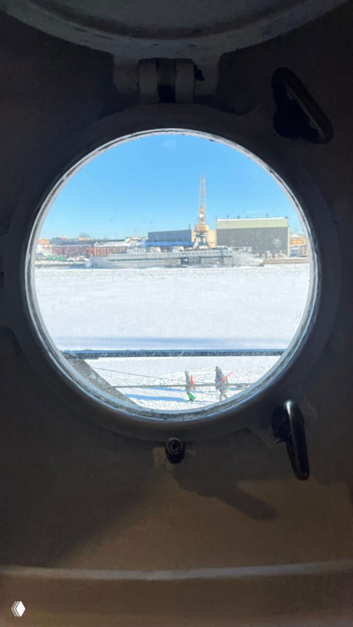 Иллюминатор ледокола с круглым проёмом, через который видна заледеневшая вода и промышленные здания верфи на другом берегу.