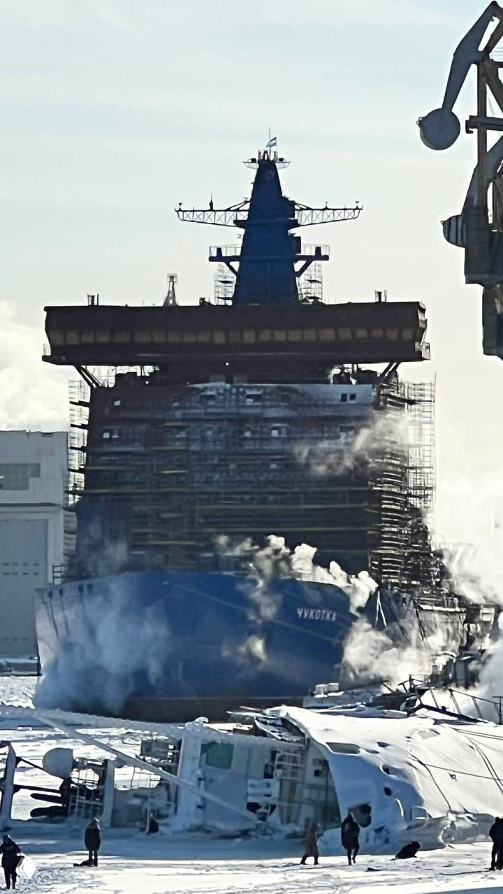 Носовая часть строящегося атомного ледокола проекта 22220 «Чукотка» в доке, строительные леса и пар над палубой на фоне верфи.