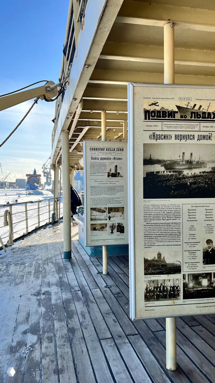 Палуба ледокола с выставочными стендами и видом на заледеневшую набережную и Адмиралтейские верфи под ясным зимним небом.