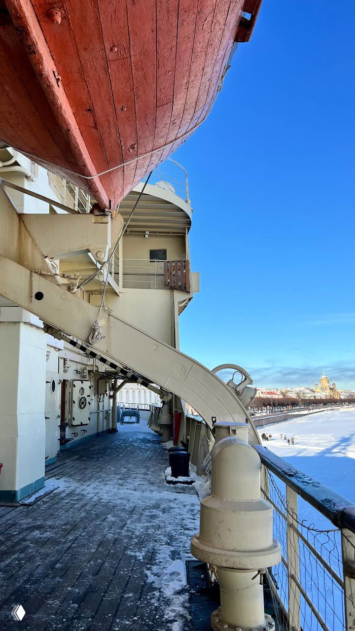 Подвешенная спасательная шлюпка на палубе ледокола, вид на конструкцию шлюпки, снежную палубу и городские постройки за бортом.