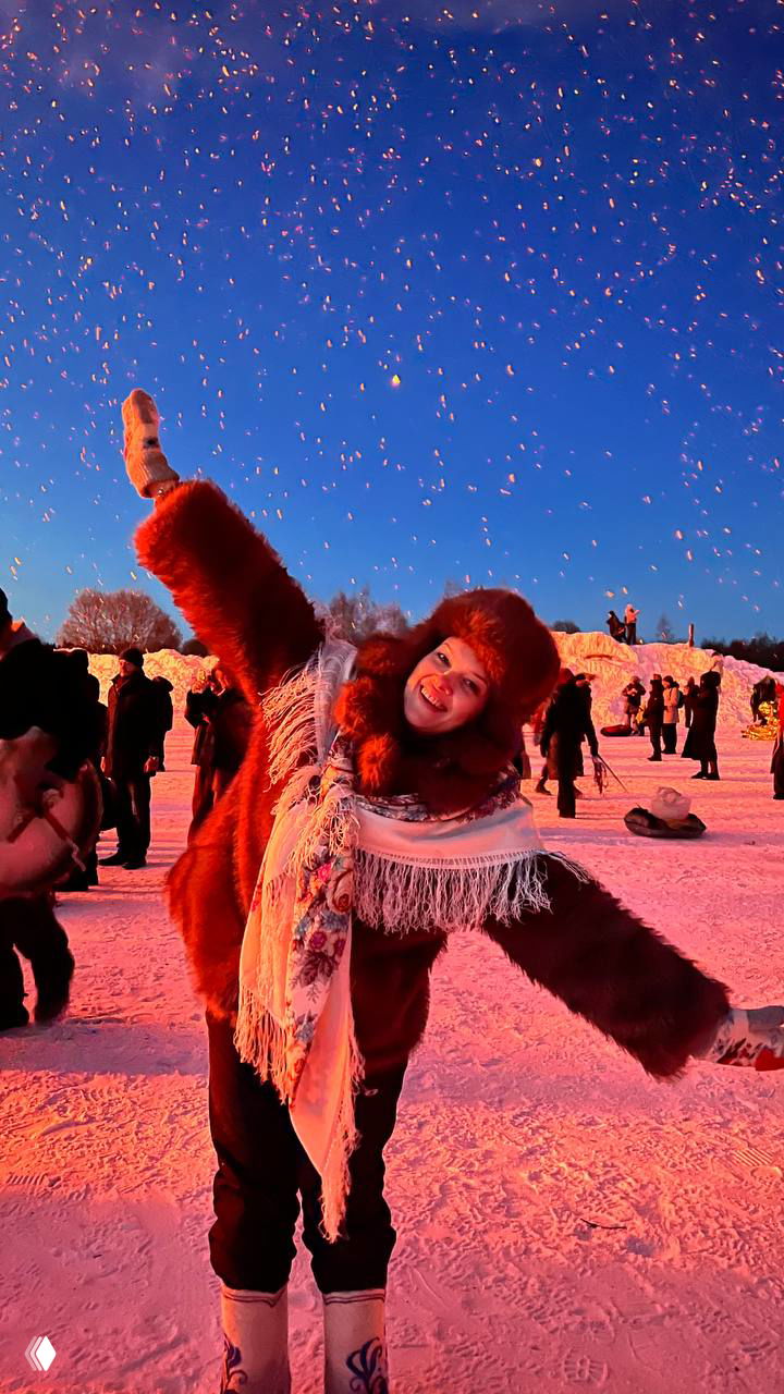 Вечерняя сцена с мощным красным светом от горящего арт-объекта: фигуры в костюмах на фоне огня, искры взлетают в небо во время сожжения «Горящего сердца» на Масленице.