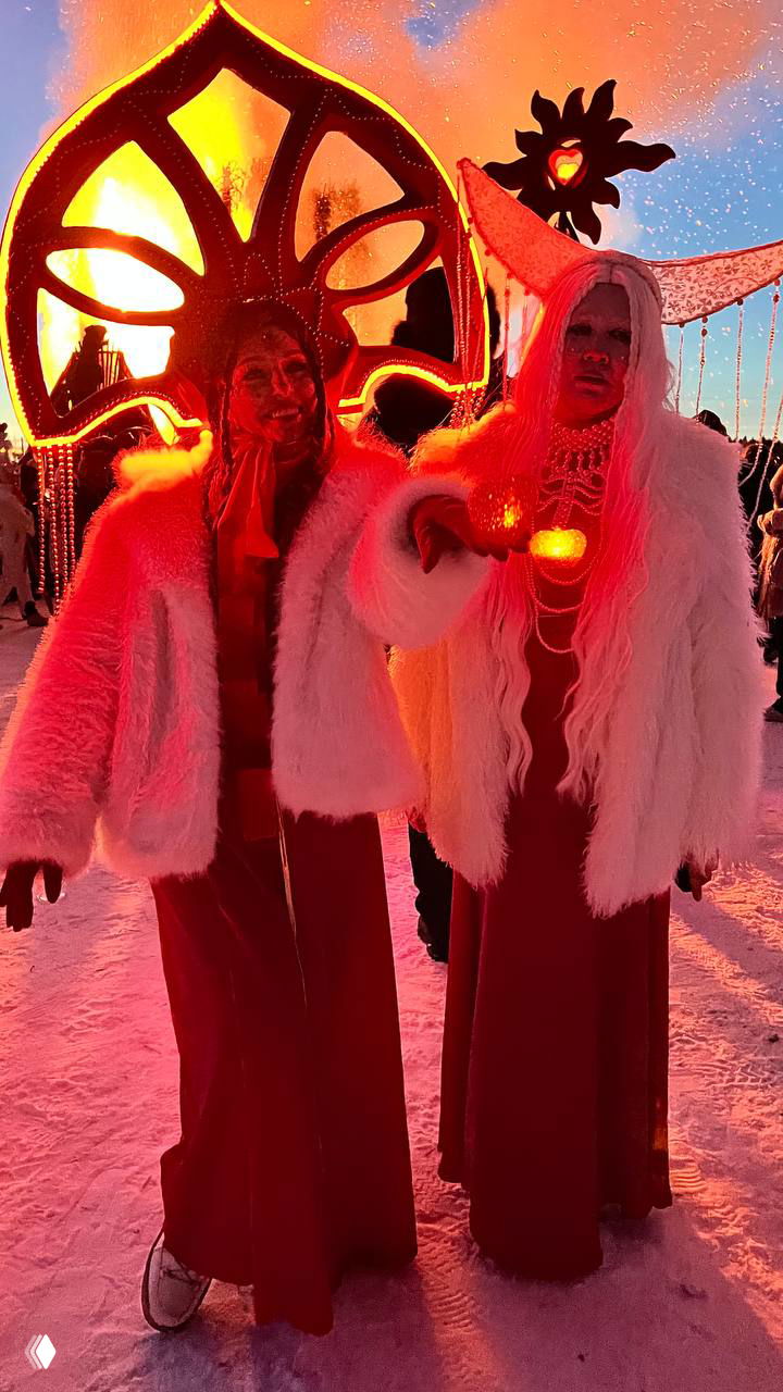 Автор в меховой шапке улыбается и держит красную ленту «Скучный Блог», позади видна деревянная лестница-инсталляция и праздничные декорации арт-парка в ясный день.