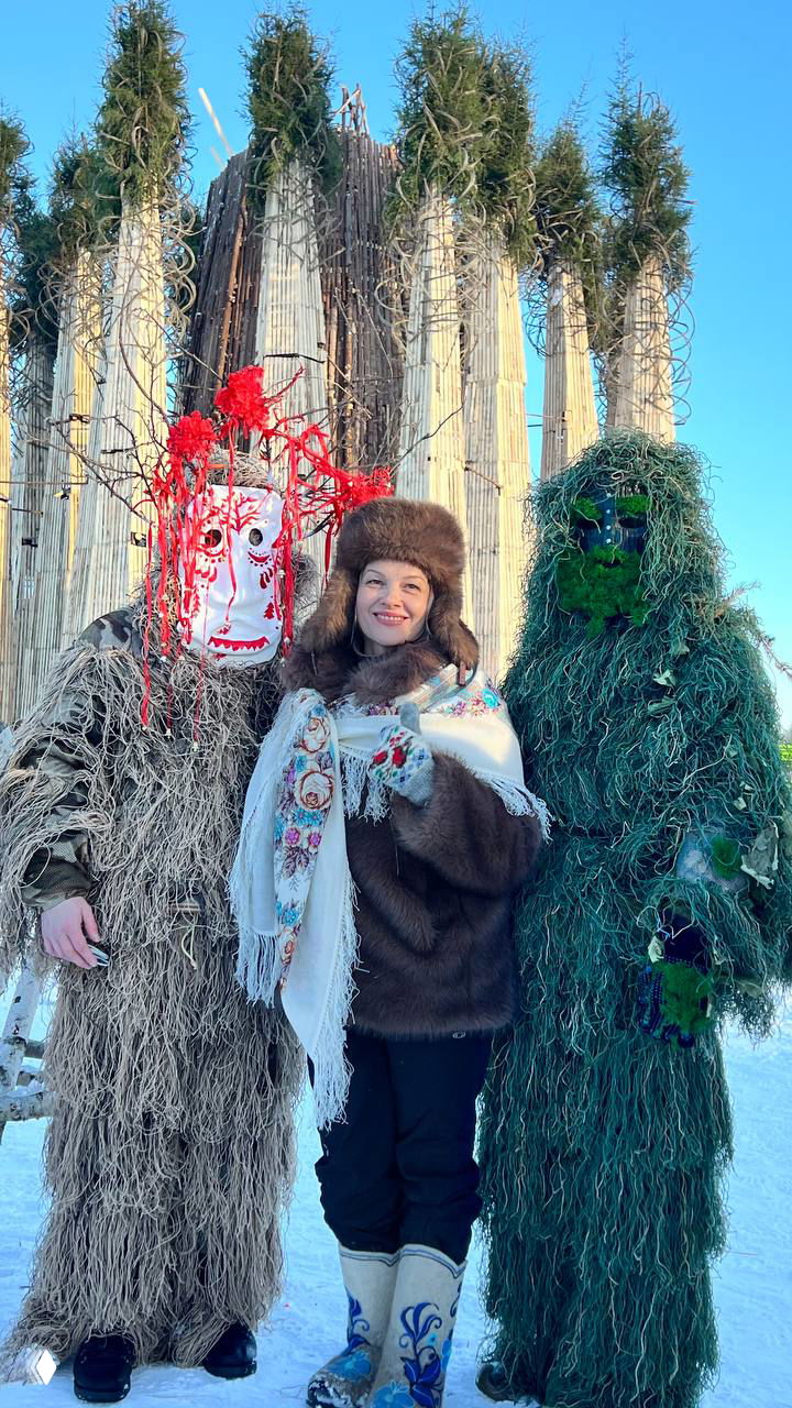 Женщина в тёплой шубе улыбается между двумя людьми в сложных масках и костюмах; позади видно крупную деревянную инсталляцию и гостей праздника в снегу.