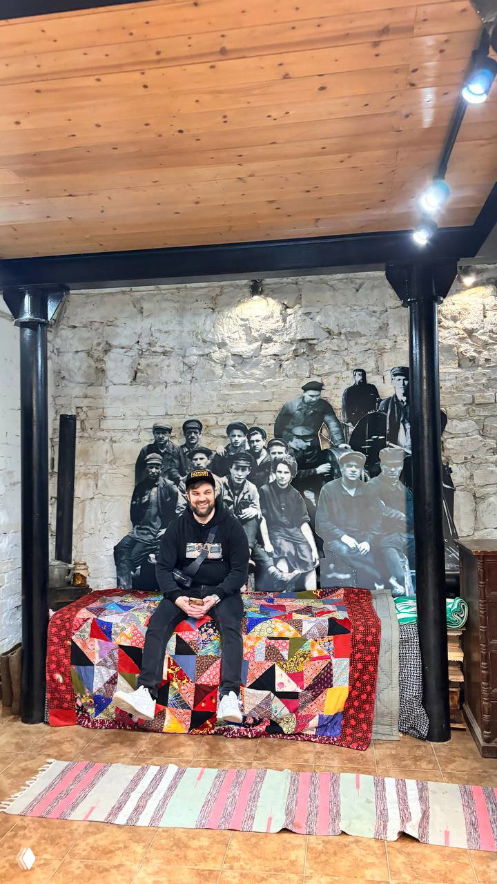 Посетитель сидит на покрытой лоскутным покрывалом лавке на фоне масштабной исторической фотографии рабочих мануфактуры на стене музея.