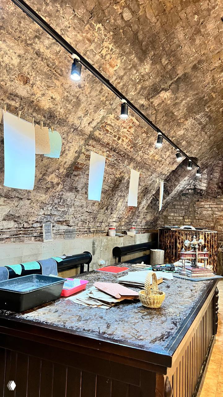 Рабочий стол мастерской: тёмная поверхность, разбросанные стопки листов, под потолком свисают листы бумаги для просушки в полукруглой каменной мастерской.