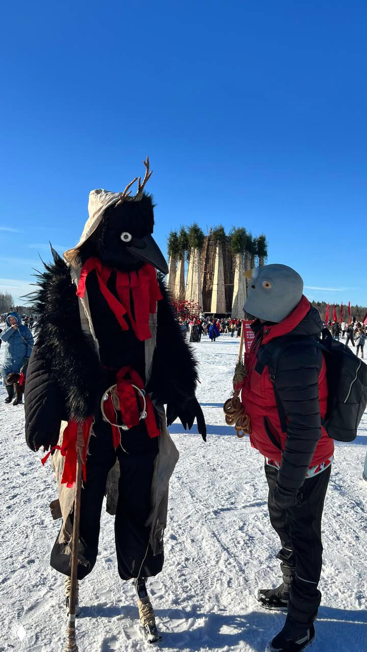 Два человека в чёрных и серых костюмах с пернатыми масками беседуют на поле перед большой деревянной структурой, зимний свет и толпа участников Масленицы на заднем плане.