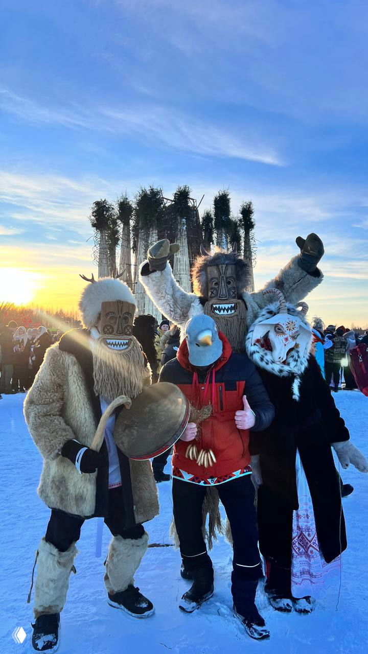Три человека в масках и традиционно стилизованных костюмах стоят перед высокой круглой деревянной инсталляцией на фоне синего неба во время Масленицы в Никола-Ленивце.