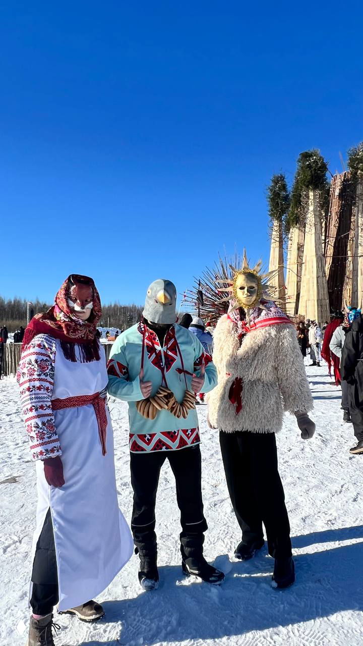 Три фигуры в разных масках и нарядах позируют вместе перед высокой круглой инсталляцией; сцена зимнего карнавала в Никола-Ленивце, солнечный день и снежная равнина.