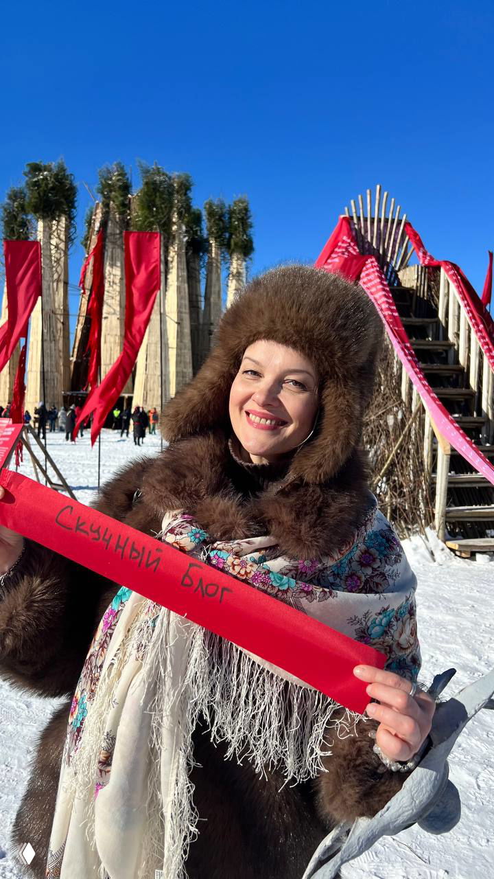 Трое людей в традиционных и оригинальных масках, один из них в солнцеподобной маске, на фоне арт-объекта и толпы — сцена карнавала во время праздника Масленицы.