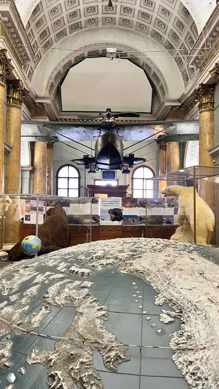 Главный зал экспозиции: макет рельефного глобуса, подвешенный полярный самолёт и чучело белого медведя на фоне колонн внутри музея.