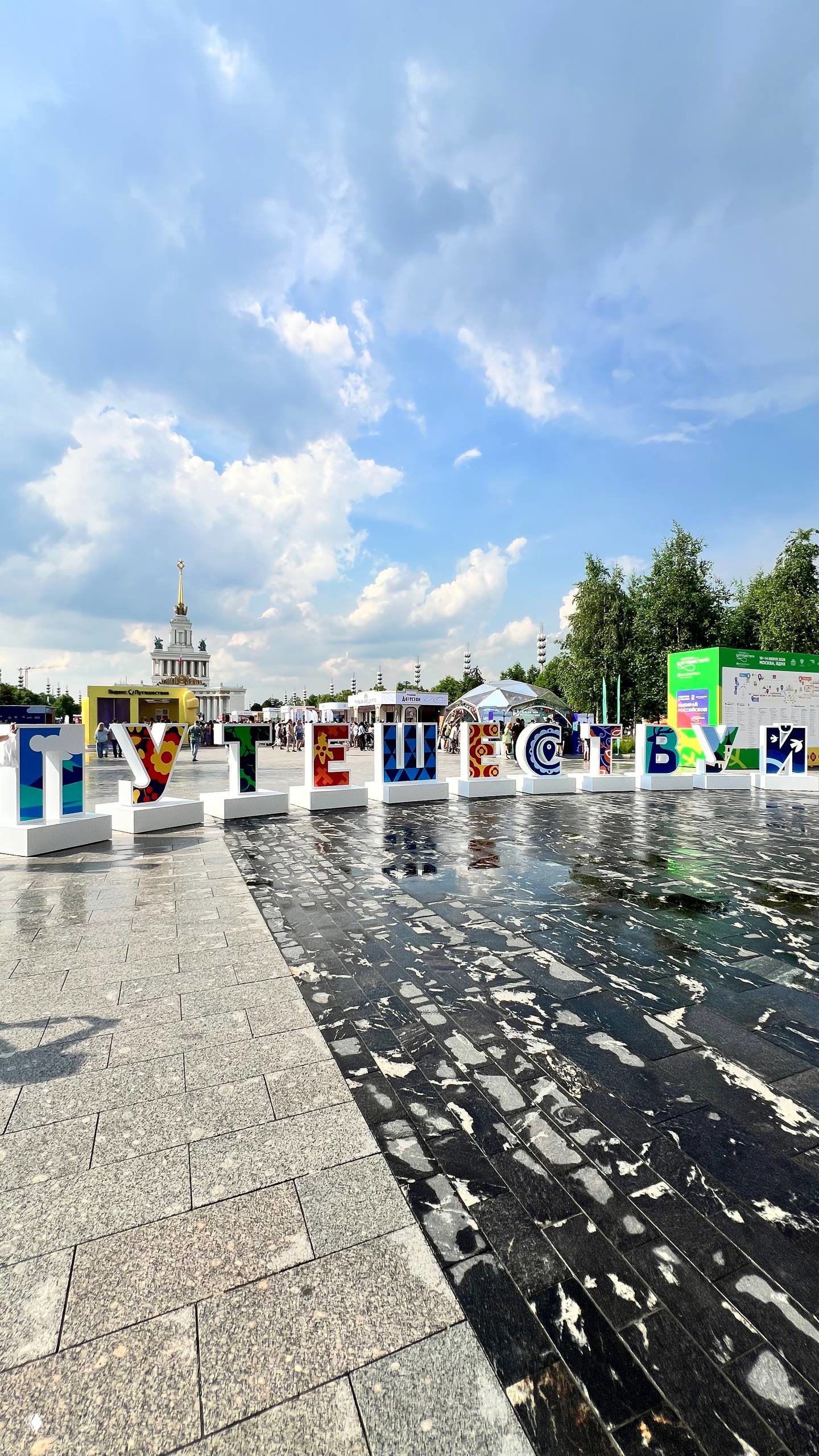 Инсталляция «ПУТЕШЕСТВУЙ» на площади ВДНХ под летним небом — большая цветная надпись и отражения на мокрой брусчатке, люди у стендов.