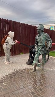 Скульптура пожарного и посетитель у памятника на мощеной площади; мокрый брусчатый тротуар и фоновые ограждения вокруг экспоната.