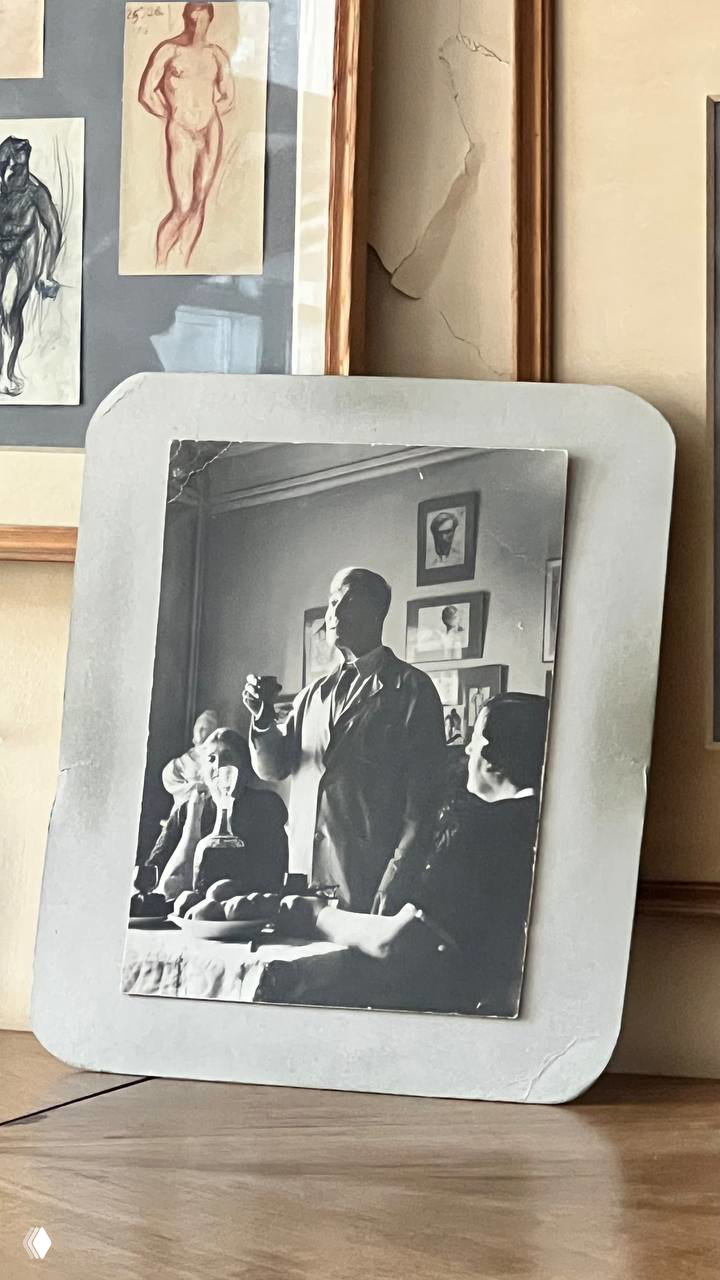 Черно-белая фотография в рамках на столе музейной комнаты: портретная сцена с людьми, рядом видны эскизы на стене и интерьер.