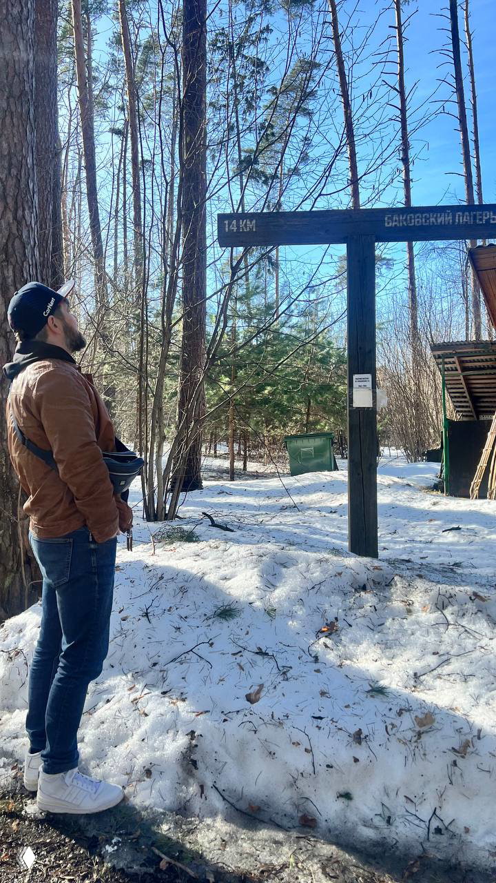 Посетитель в кепке стоит у деревянного указателя на лесной тропе в Переделкине; снег под ногами, сосны и яркое небо создают атмосферу прогулки.