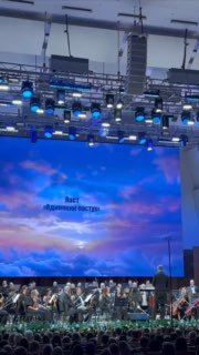 Широкий кадр сцены и экрана, на котором идёт видеоряд с изображением неба и самолётов; внизу виден оркестр и освещение зала.