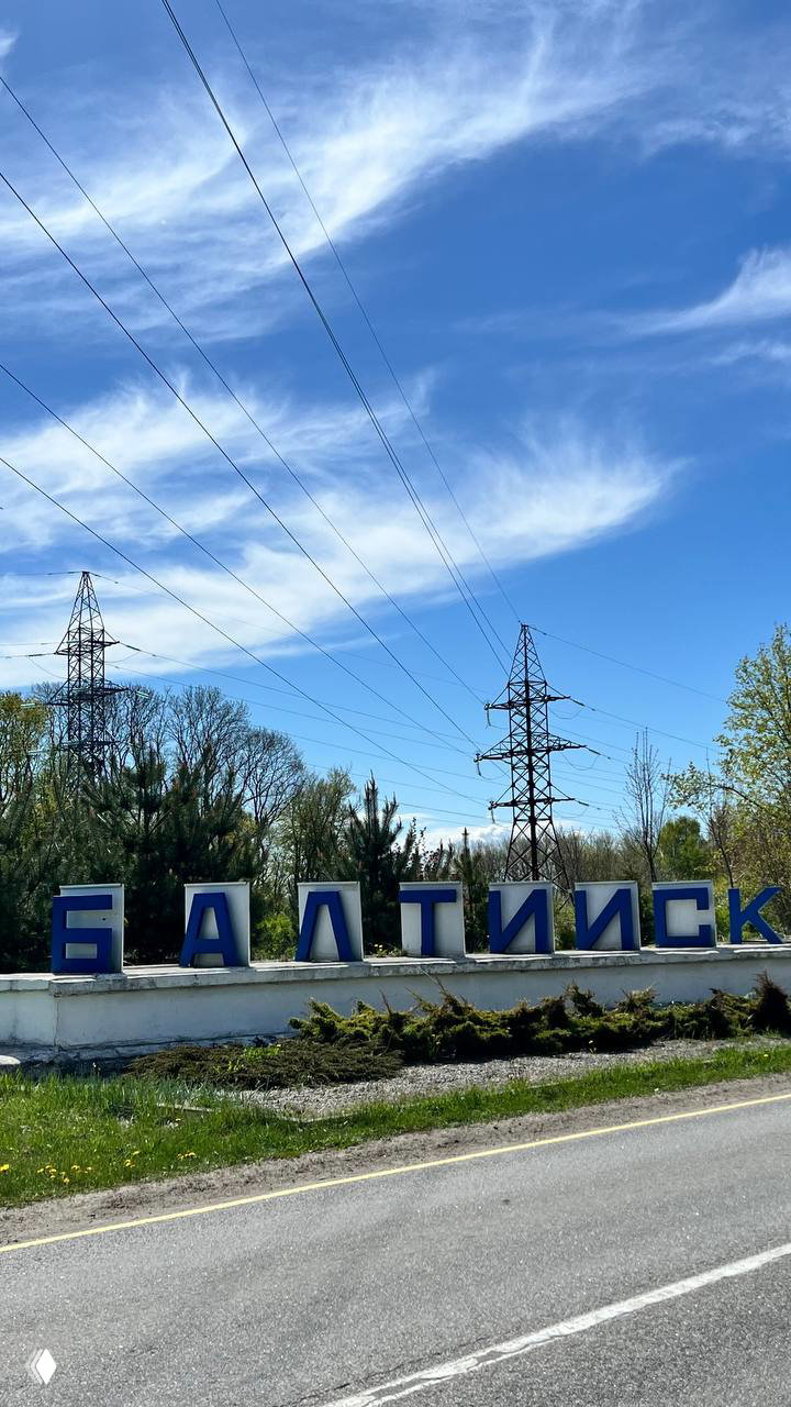Дорожный указатель и большой голубой буквенный знак «БАЛТИЙСК» у въезда в город на фоне линий электропередач и весеннего неба, вид с трассы.