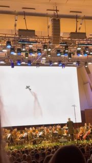 Кадр сценического видеоряда с силуэтом пролетающего самолёта над оркестром; сочетание музыкальной сцены и авиационных образов.