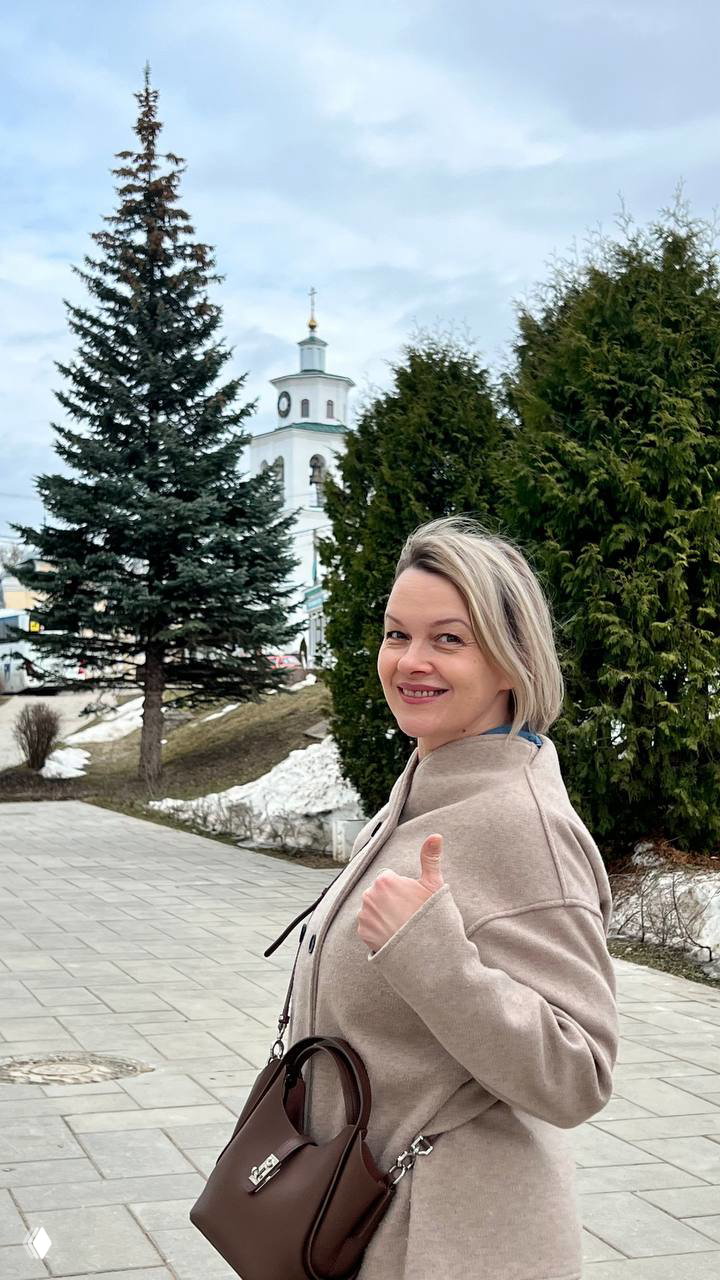 Женщина улыбается и показывает палец вверх на фоне ели и церкви в посёлке Полотняный Завод; прогулочная сцена без узнаваемых личных данных