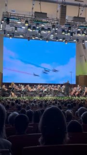 Ещё один фрагмент с экрана и сценой: фигуры высшего пилотажа в видеоролике и музыканты оркестра внизу кадра.