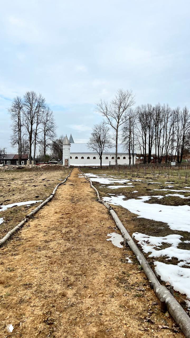 Дорожка по территории парка Полотняного Завода, остатки снега по краям и белая длинная постройка вдалеке — ранняя весна в усадьбе