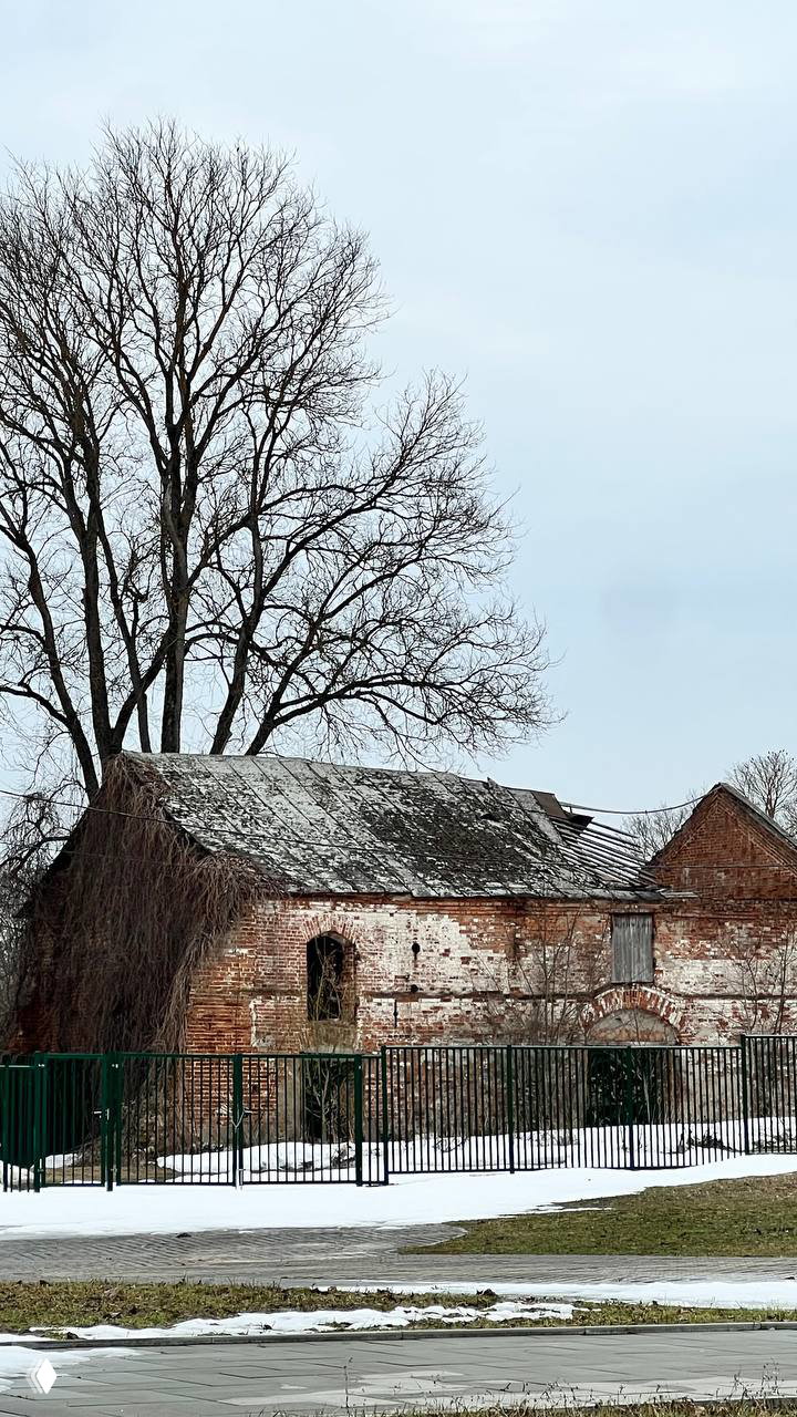 Старая кирпичная хозяйственная постройка с частично разрушенной кровлей и обвалившейся кладкой на фоне голых деревьев и снега