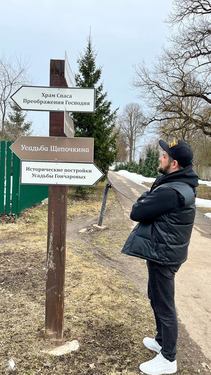 Указатель и человек, рассматривающий направление к «Усадьбе Щепочкина» и храму Спаса Преображения — сценка ориентирования в усадебном парке