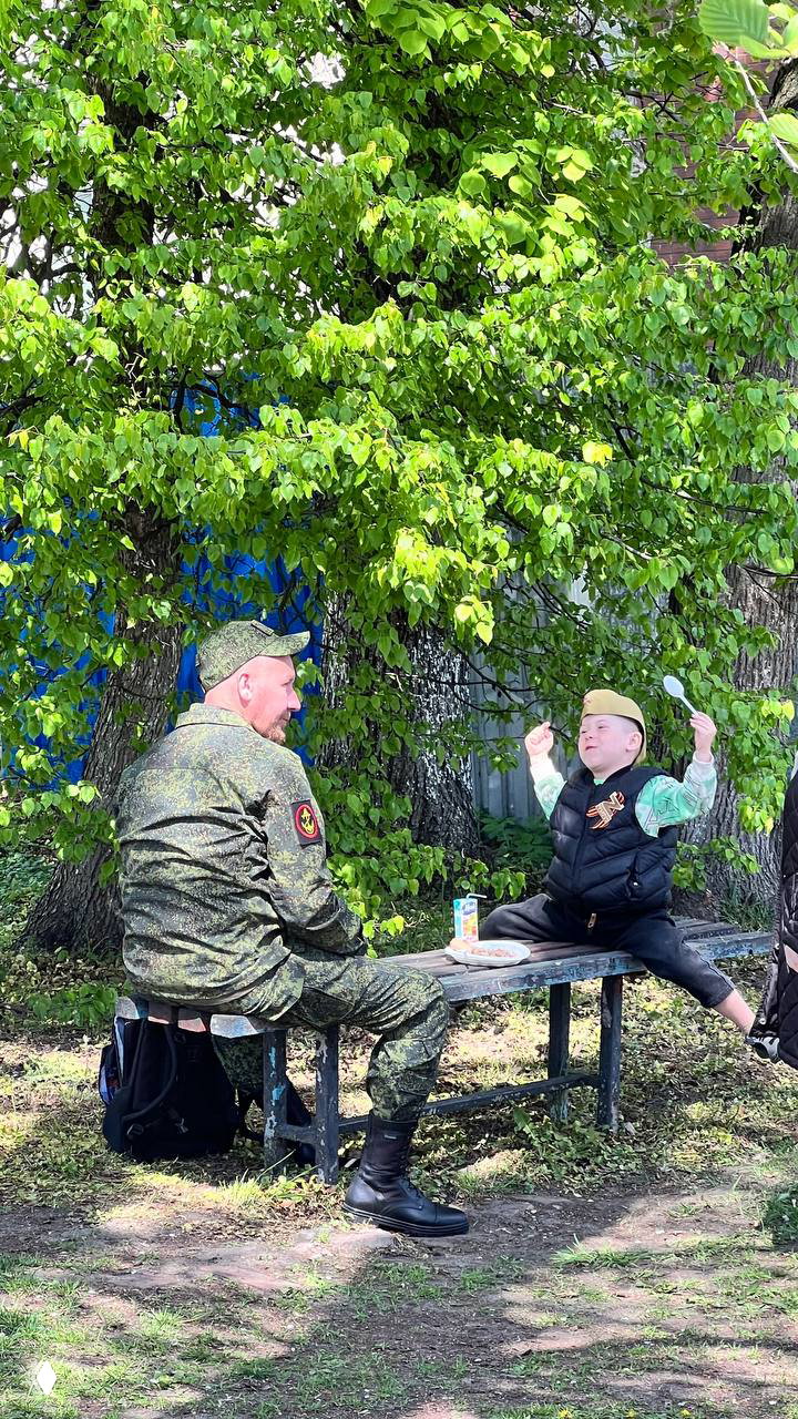 Старая скамья под деревом, рядом мужчина в военной форме и ребёнок в пилотке с ложкой — семейная сцена на зелёной поляне в честь 9 Мая, тихий повседневный момент.