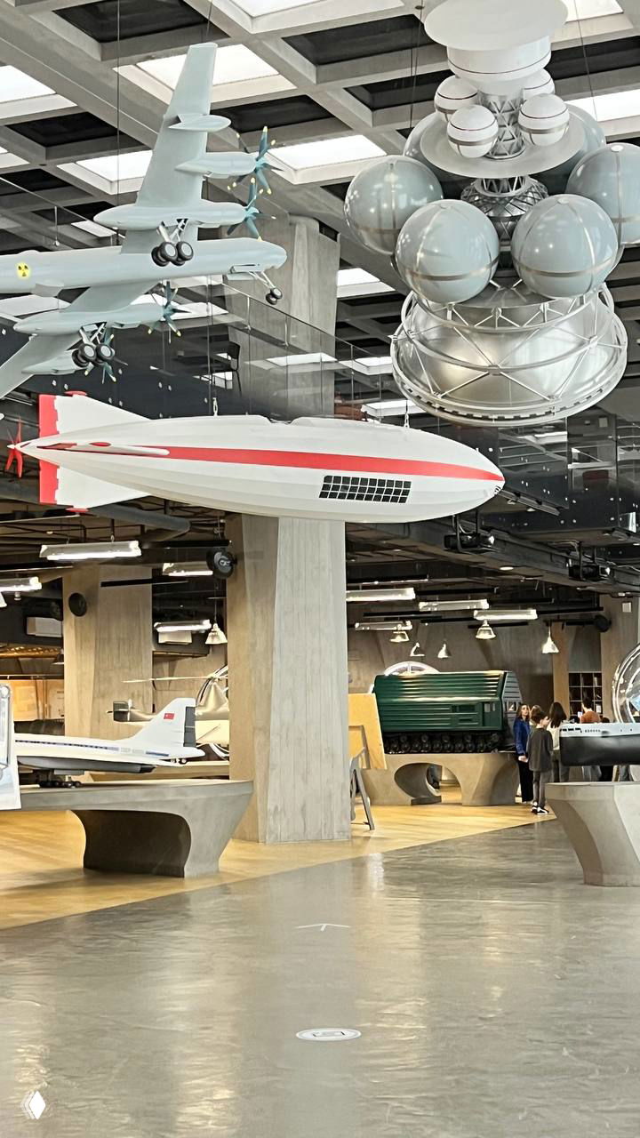 Подвешенные модели самолёта и дирижабля, а также сферические элементы в большом выставочном зале музея «Атом», вид на экспонаты и стойки экспозиции.