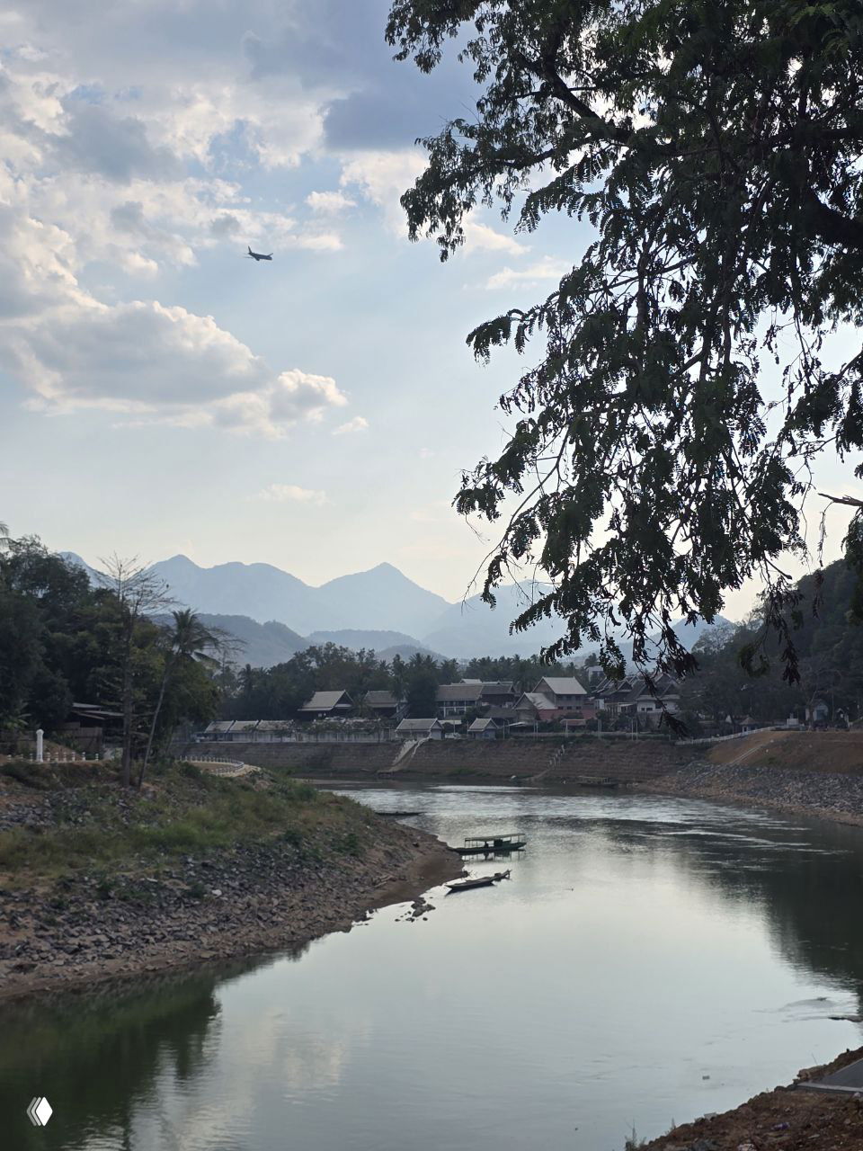 Река Меконг с небольшими лодками у берега, отражения воды и силуэты гор на горизонте под облачным небом, в небе виден пролетающий самолёт.