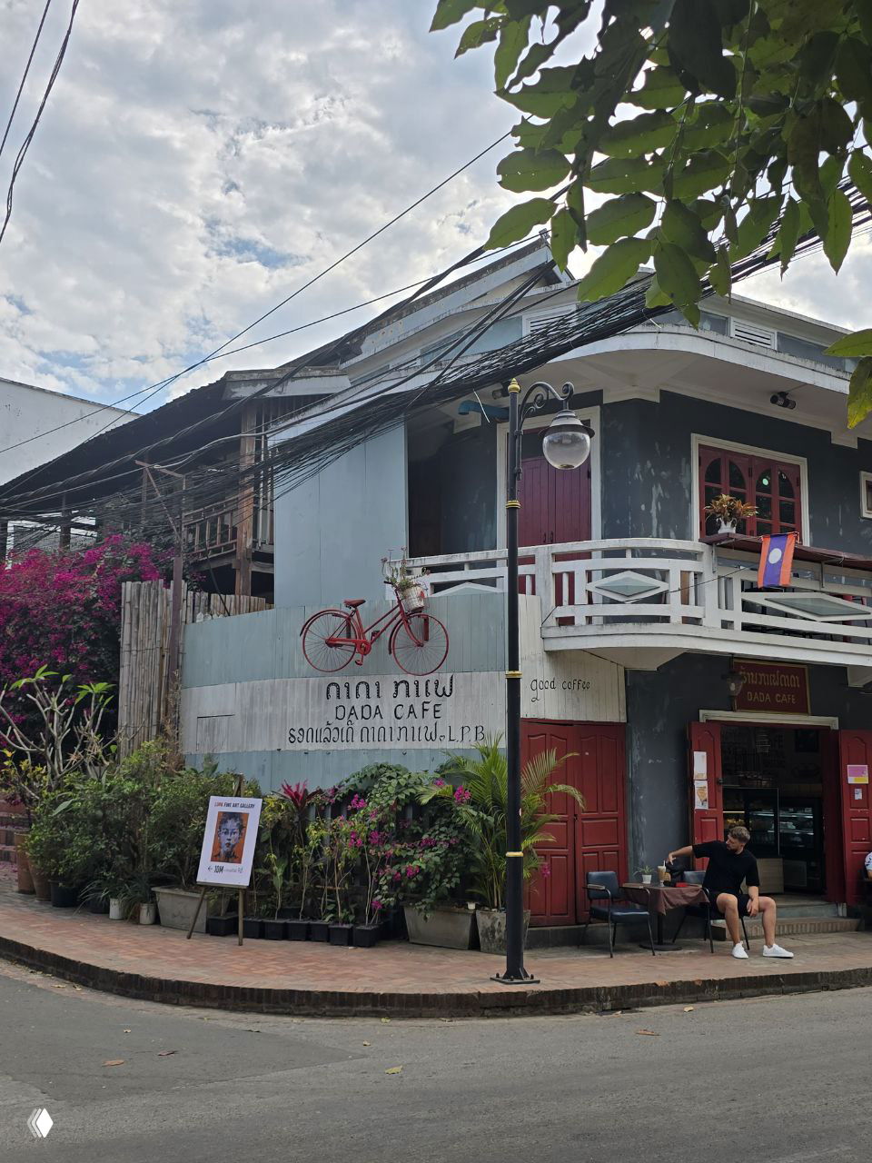 Фасад уютного Dada Cafe с декоративным красным велосипедом на вывеске, растениями в кадке и креслами у входа на тихой улице старого города Луанг Прабанга.