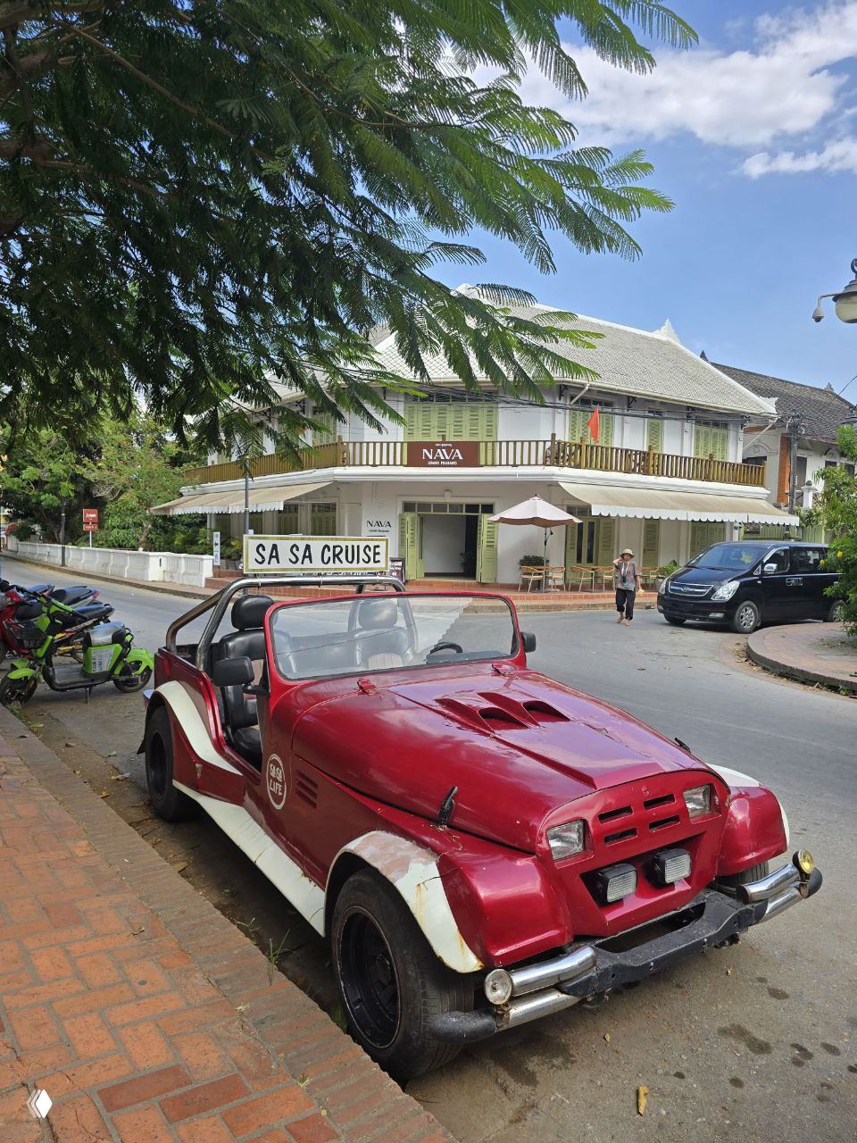 Красный ретро-кабриолет припаркован на кирпичной брусчатке рядом с улицей и кафе, создавая туристический колорит и фотогеничный стрит-вид города.