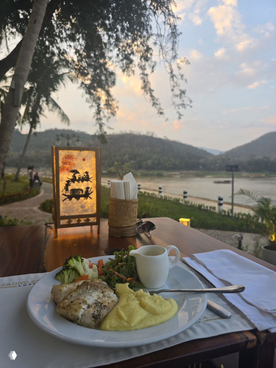 Ужин на открытой веранде у реки: тарелка с рыбой и пюре, бокал и сервировка на фоне реки Меконг и холмов при мягком закатном свете, атмосферный столик с видом.