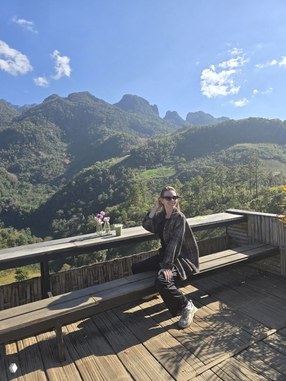 Девушка в солнцезащитных очках сидит на деревянной веранде с видом на глубокие зелёные горы и яркое солнечное небо Чиангмая.