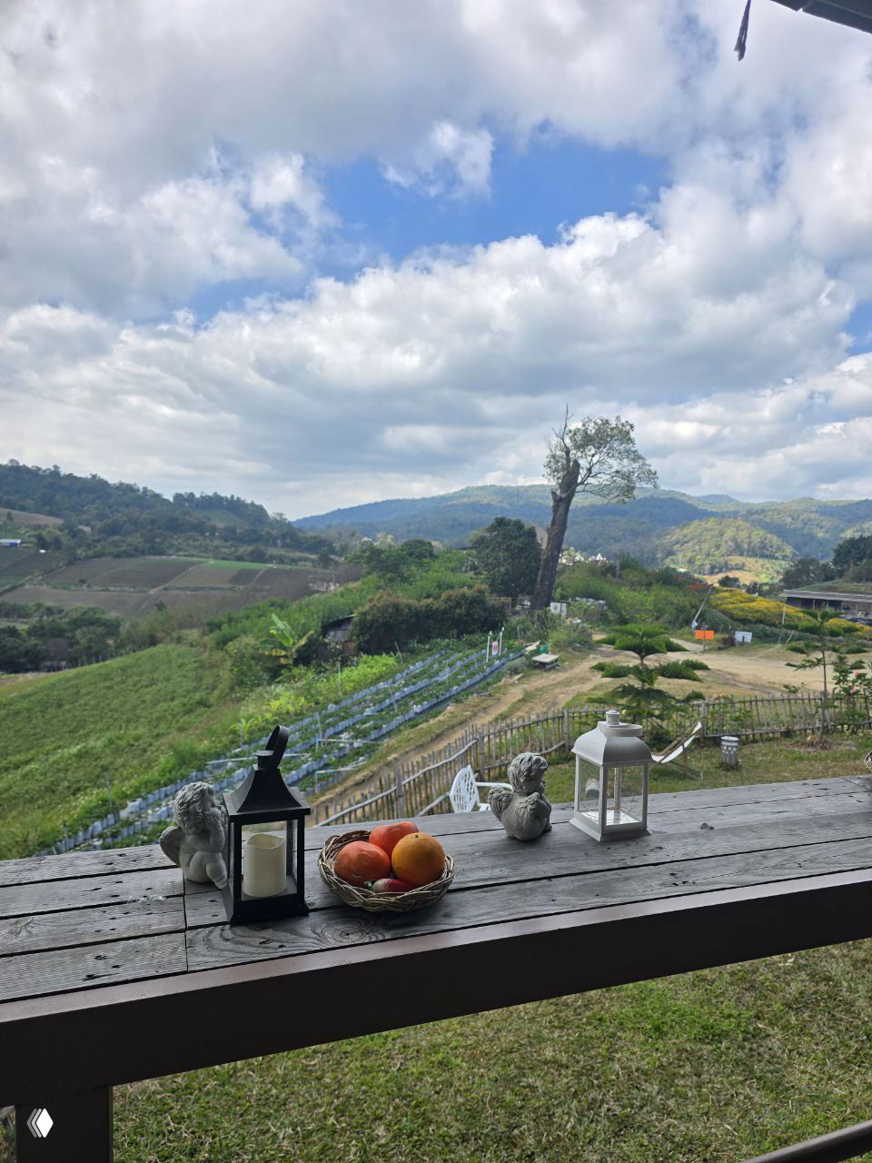 Вид с террасы на горные поля и фермы Чиангмая: на столе корзина с фруктами, свечи и декоративные фигурки на фоне холмов и облачного неба.