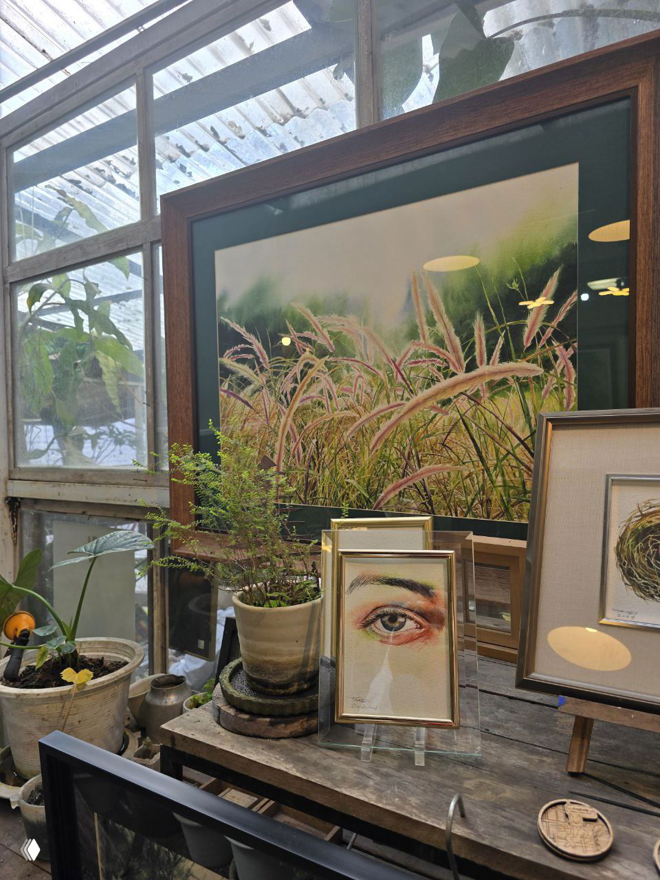 Интерьер небольшого арт-кафе или студии: картины в рамках, растения в горшках и творческая композиция на деревянном столе в тёплом свете.