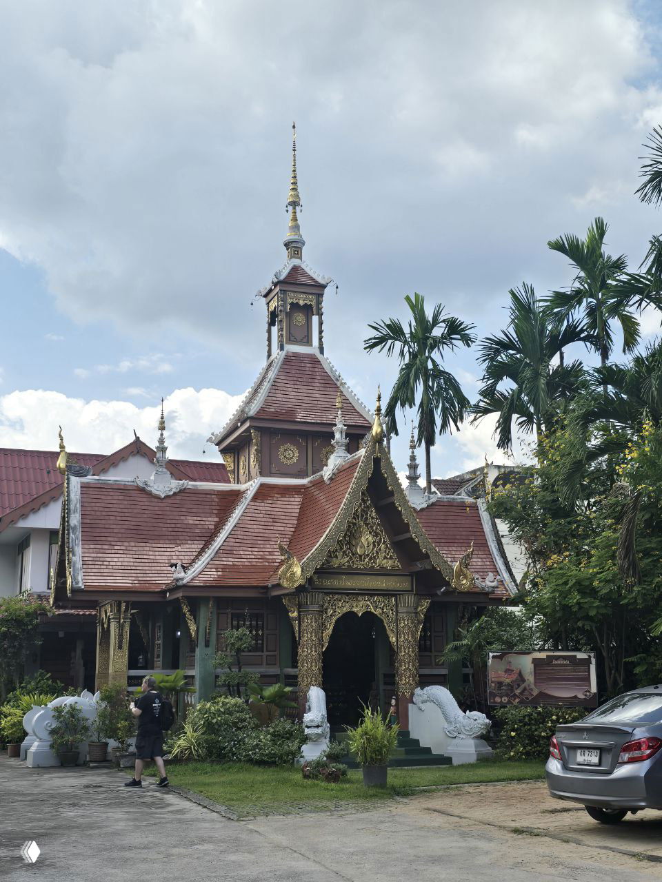 Традиционное тайское здание с резным фасадом и золотыми элементами у входа, окружённое тропическими растениями и уходящими в небо облаками.