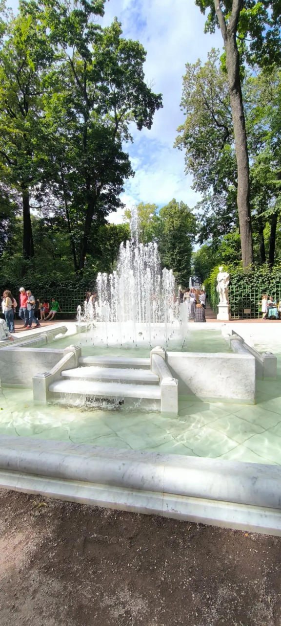 Вертикальная фотография паркового фонтана на тёплый день: фонтан в центре, вокруг зелёные деревья и люди, сцена спокойного отдыха — метафора паузы и скуки.