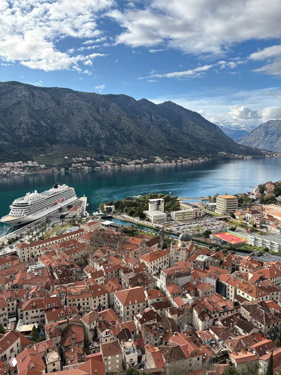 Прибрежный город с красными крышами, пришвартованный круизный лайнер и глубокий залив на фоне гор — вид сверху из отпуска.