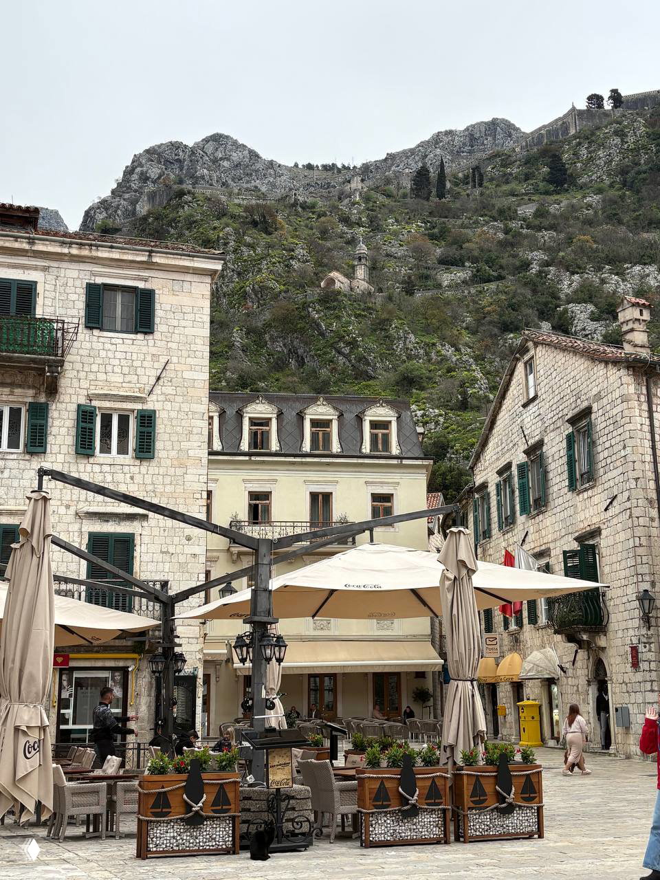 Каменная городская площадь с уличными зонтиками и столиками кафе у подножия крутого скалистого холма — уютный городской кадр.