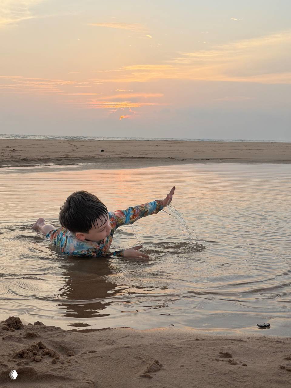 Мальчик плескается в мелкой воде на фоне закатного неба, расплескивает воду и наслаждается простыми пляжными играми без правил.